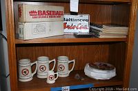 Shelf containing two Fleer Baseball trading card boxes, four ceramic mugs with baseball logos, a round white baseball glove display case, a Baltimore Orioles decorative plate, and a stack of baseball cards or similar paper items.
