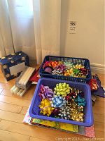 Two blue plastic baskets filled with a variety of colorful gift bows and curling ribbons, 13 white taper candles in packaging, sun and moon recipe box, and gift bags stacked on the floor.