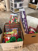 Box containing various rolls of Christmas wrapping paper, gift boxes, bags, ribbons and bows arranged on floor.