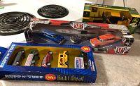Photo showing ERTL John Deere Road Grader in yellow and black with original John Deere themed box, M2 Die Cast models of 1957 Chevy Bel Air cars in blue, black and orange/red colors, and five Ruff 'n' Tuff toy cars and trucks in various colors all sealed in original packaging.