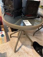 Small round patio table with glass top and beige metal frame, shown with two black heater units on top and some nearby household items, carpeted floor visible.