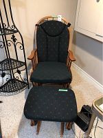 Front view of wood frame gliding rocker chair with high back and matching ottoman, both with dark patterned cushions.
