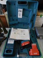 Blue plastic hard carrying case with Makita cordless drill accessories and user manual inside, case open and contents displayed, showing charger, two batteries, and paperwork.