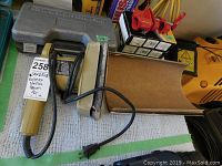 Photo showing electric sander, rubber sanding drum case, and two red pipe clamps in boxes on table.