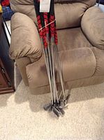 Full view of seven golf irons leaning against a chair showing the shafts with red and black grips and metal club heads.