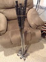 Nine Select Model golf irons resting upright indoors on a beige armchair, grips secured with a rubber band.