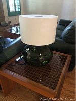 Green glass table lamp with white drum shade positioned on wooden table with grid pattern top in living room setting with black leather furniture.