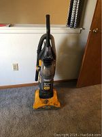 Full view of yellow Eureka upright vacuum cleaner standing on carpeted floor, showing body, handle, and hose attachment.