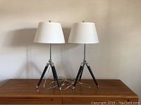 Front view of both table lamps on wooden surface with white shades and tripod bases, showing overall design and height.