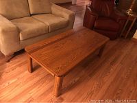 Rectangular oak coffee table with natural wood grain finish, shown beside beige sofa and red chair on hardwood floor.