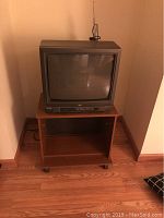 Front view of the CRT television on the wooden rolling stand showing TV screen and VHS slot.