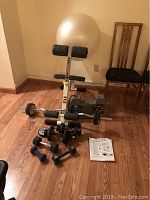 White and black adjustable weight bench with exercise ball behind, free weights in front including dumbbells and barbell.