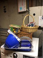 Assortment showing stacked picnic supplies including blue and green plastic bowls, disposable aluminum pans, plastic utensils and napkins in sealed packaging, and woven baskets.