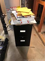 Black metal 2-drawer file cabinet with top covered by various mailing supplies including yellow padded envelopes and FedEx shipping box.