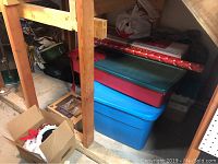 Photo of stacked plastic bins, wrapping paper, and cardboard box with decorations under a wooden structure