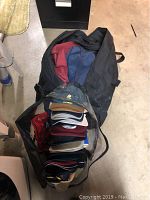 Top view of a large black duffel bag fully packed with about a dozen baseball hats stacked one on top of another, showing colors like red, blue, black, white, and navy.