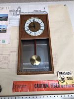 Full view of the wooden wall clock showing the clock face with Roman numerals and the pendulum behind a glass panel.