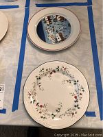 Two decorative plates displayed on a table marked with blue tape lines, showing one with a floral pattern and the other with a central house and bird design.