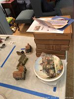 Stack of boxes with plates on top showing Country Cottage plate art. Also shows other small cottage collectible figurines nearby.
