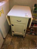 Front view of an off-white vintage drop leaf cabinet with metal top and one drawer, showing condition and surroundings.