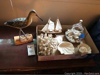 Wide shot showing wooden bird sculpture, model sailboat, white decorative coral, glass jar with shells, and several ceramic shells arranged in cardboard box.