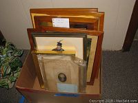 Box filled with various framed and matted prints and lithographs of different sizes, seen stacked upright and resting on side in cardboard container.