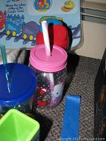 Photo of placemat in the background, with multiple sippy cups including pink mason jar style cup with straw, blue cup with straw, and green juice holder, on carpeted floor