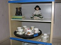 Shelf showing porcelain doll, vintage binoculars on books, and Lazy Susan with tea cups and saucers.