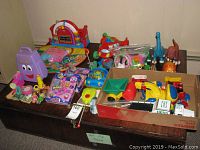 Photo showing the assortment of children's toys including Fisher-Price playmat, boxed Shopkins toys, toy trucks, dinosaur figures, and other miscellaneous toys on a table.