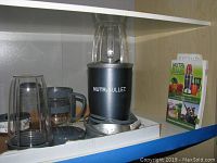 NutriBullet blender base with clear blending cup attached, additional cup with handle and lid, user guide and recipe book visible on shelf.