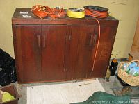 Front view of dark brown wooden storage cabinet with two doors, visible wear, items on top including electrical cords.