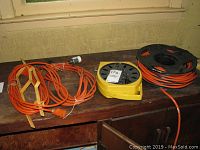 Four orange extension cords on a wooden surface, including one on yellow cord storage wheel and one black and yellow Multiplug Reel