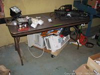 Folding table with various items placed on top in a garage setting. Table surface shows wear including dings, scratches and water damage.