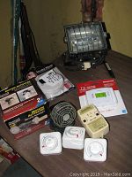 Photo shows multiple items including Regent Security motion flood light with protective grille, Code One smoke alarm in packaging, three Intermatic timers, small mini fan, Guardian circuit interrupter, and Honeywell thermostat in packaging.