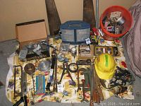 Overview of all hand tools, fasteners, safety gear spread on the floor including toolbox, safety hat, wires, nails, clamps, hammer, wrenches, and various hardware.