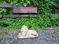Wide view showing the wooden American flag mounted on a fence above the lion statue on gravel ground with greenery in background.