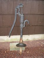 Full view of vintage iron water pump installed outdoors against a wood siding wall with rust and weathering on metal surfaces.