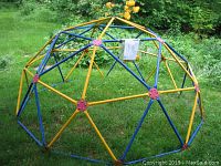 Full view of geodesic dome climber with alternating yellow and blue metal bars and red hex bolt connectors on grass in outdoor setting.