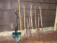 Full view of all garden tools including shovels, rakes, pitchforks, axe, pick and snow shovel aligned and leaning against a wooden wall outside.