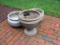 Photo showing the three outdoor planters on a brick patio with green grass background. Two smaller plastic planters with light wear and one larger cracked cement planter with decorative beaded rim and pedestal base.