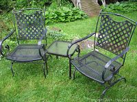 Set of two black metal rocking chairs and one black metal side table with glass top displayed on grass outside.