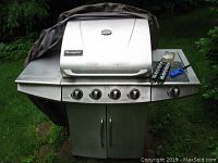 Charmglow stainless steel outdoor propane grill with attached propane tank, grills, control knobs, torn cover, grilling accessories on right side shelf.