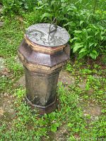 Full view of an antique chimney pot standing on garden ground surrounded by green plants. The pot is octagonal, metal with weathering and rust marks, with a sundial mounted on top.