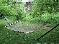Full view of rope hammock suspended on metal frame outdoors among trees and grass.
