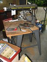 Radial arm saw on metal stand with wooden table, multiple blades and manuals visible, vacuum hose attached at rear.