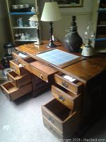 Photo showing 8 drawer wooden desk with some drawers open, desk surface with lamp and notebook. Desk is medium-tone wood with metal handles.