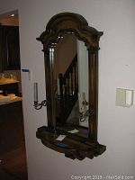 Front diagonal view of the wall mirror with wooden scalloped frame, shelf at bottom, and metal candle holders on sides.