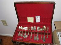 Wide view of open wooden chest with red-velvet lining showing scattered sterling flatware pieces.