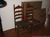 Two antique wooden rushed chairs side by side, one with a dark rush seat and one with a light rush seat. Ladder back design with wavy horizontal slats.