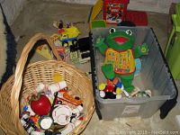 View showing large wicker basket filled with toy tea set items and other small toys alongside a large plastic tub holding a green stuffed frog toy, alphabet toy, and small toys.